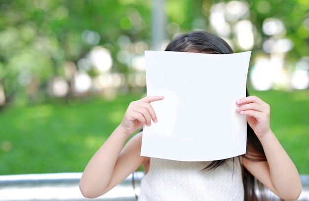 Cara de ocultación de la niña pequeña detrás del Libro Blanco en blanco en el jardín verde.