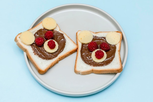 Cara de oso divertido pan tostado sándwich con mantequilla de maní, queso y frambuesa sobre fondo azul placa. Niños niño dulce postre desayuno almuerzo