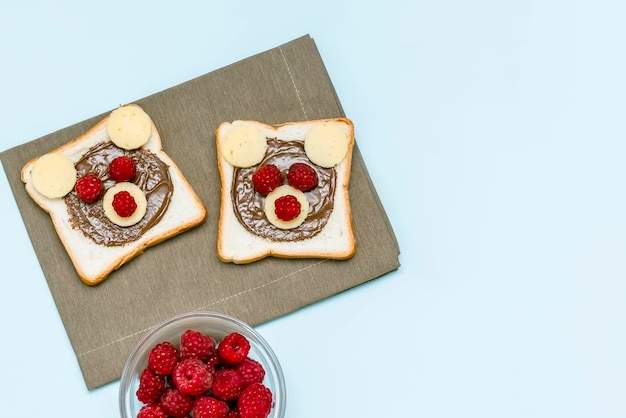 Cara de oso divertido pan tostado sándwich con mantequilla de maní, queso y frambuesa sobre fondo azul servilleta. Niños niño postre dulce desayuno almuerzo comida cerrar