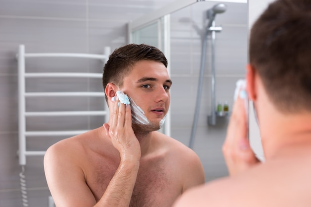 Cara passando gel de barbear no rosto em frente ao espelho no banheiro moderno de azulejos de casa