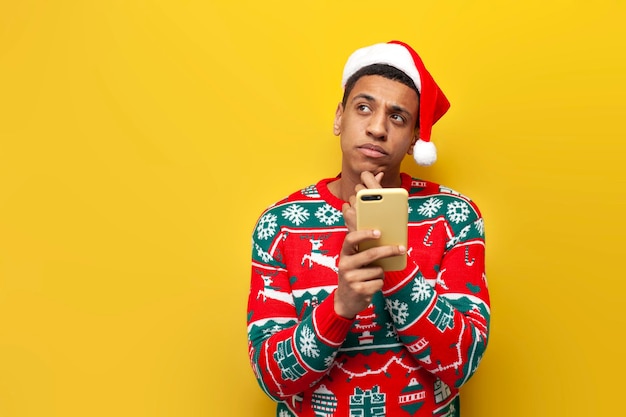 cara pensativo afro-americano com roupas de ano novo e chapéu de Papai Noel usa smartphone e pensa