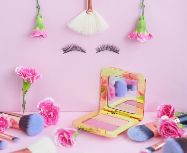 Cara con pestañas postizas y flores, pinceles de maquillaje de primera calidad sobre un fondo de color rosa, cosméticos creativos planos