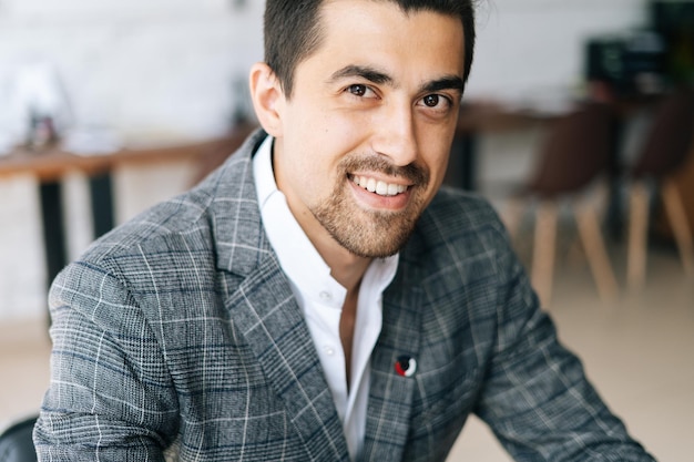 Cara de primer plano de un alegre y elegante hombre de negocios con traje de moda sentado en un escritorio en una moderna sala de oficinas mirando a la cámara