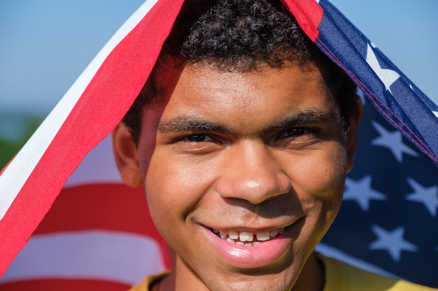 Foto cara de primer plano del hombre afroamericano feliz se envuelve en la bandera de los estados unidos y mira a la cámara y sonríe al aire libre en verano