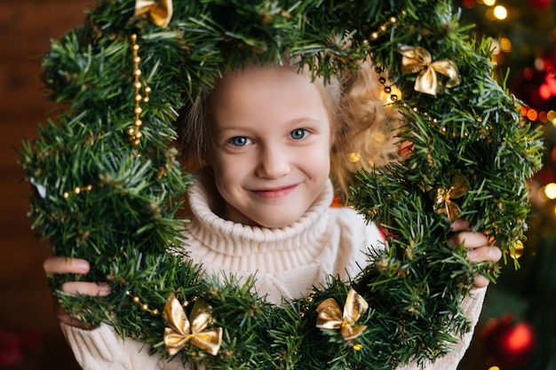 Cara de primer plano de una niña pequeña rubia feliz sosteniendo una corona redonda de Navidad en la cara de pie en una habitación oscura y acogedora en el tiempo de Navidad