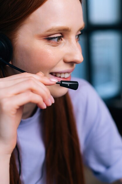 Cara de primer plano del operador alegre joven con auriculares durante la atención al cliente en la oficina en casa.