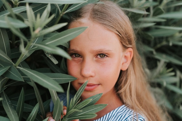 Cara de retrato de una niña pequeña feliz y sincera de ocho años con cabello largo y rubio y ojos verdes en el fondo de plantas verdes durante un concepto de salud mental de la generación z de viajes de vacaciones de verano