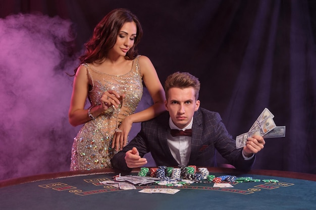 Cara segurando cash play poker no cassino sentado à mesa com pilhas de cartões de fichas comemorando a vitória com fundo de fumaça preta feminina Closeup