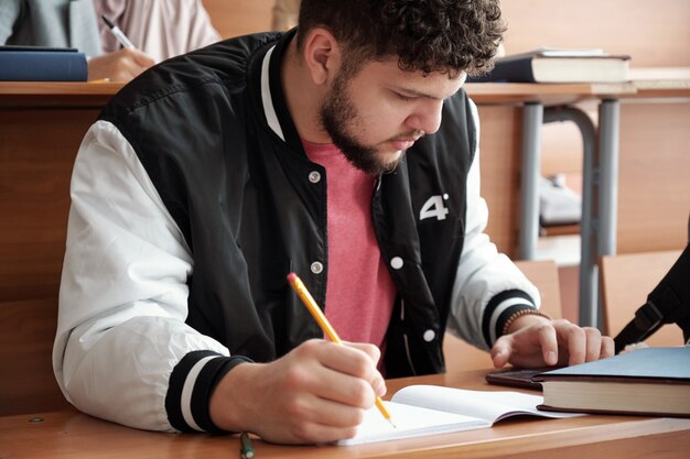 Cara sério em roupas casuais curvando-se sobre a mesa e fazendo anotações no caderno enquanto rola a tela no smartphone durante uma palestra na universidade