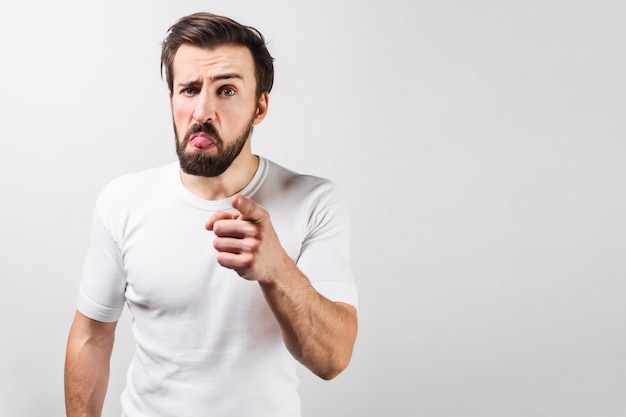 Cara sério na camisa branca está de pé no lado esquerdo de um quadro e apontando para a frente. Seu rosto mostra algumas emoções estranhas. Cortar vista. Isolado na parede branca