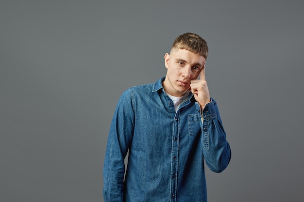 Cara sério, vestido com uma camisa jeans em pé com a mão no rosto no estúdio sobre o fundo cinza.