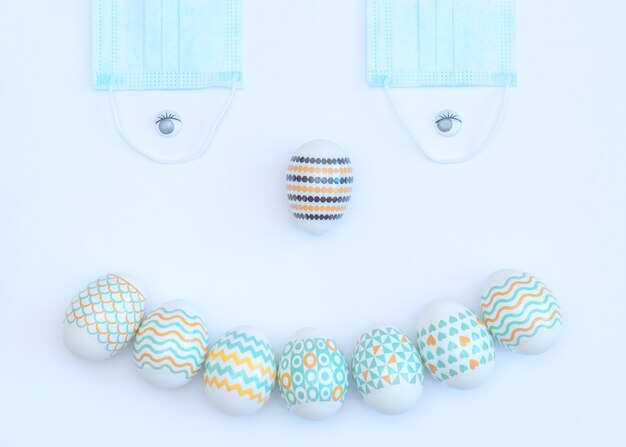 Cara sonriente hecha de huevos de Pascua decorados y mascarillas médicas en blanco