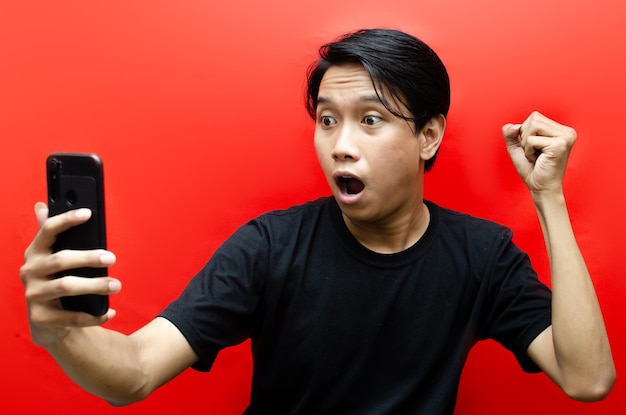 Cara sorprendida de un hombre asiático con camisa negra de fondo rojo. hombre sorprendido mientras sostiene el teléfono