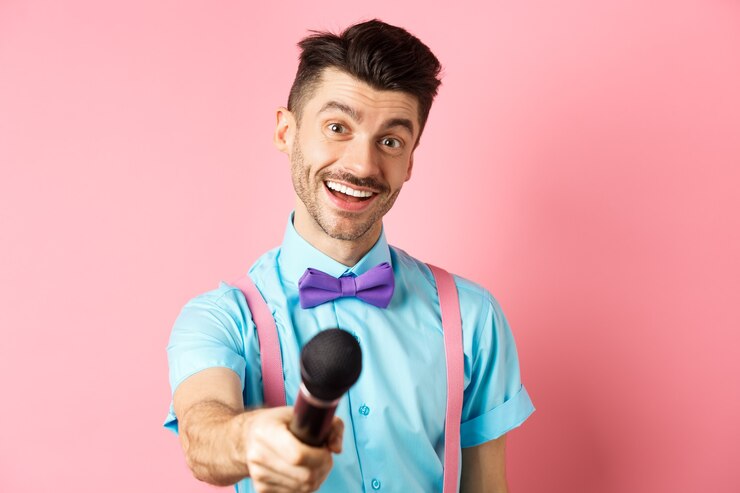 Foto cara sorridente com bigode e gravata borboleta pedindo comentários ou entrevista, estique a mão com o microfone, de pé no fundo rosa.