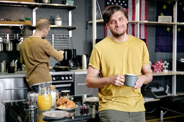 Cara sorridente em pé na cozinha e tomando uma bebida, o marido cozinhando o café da manhã no fundo