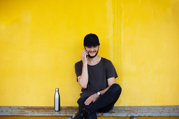 Cara sorridente está sentado no amarelo perto da garrafa térmica de aço e falando no smartphone.