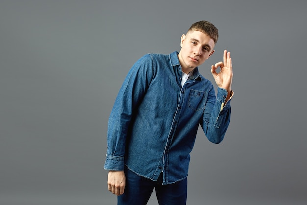 Cara sorridente vestido com uma camisa jeans mostra com os dedos ok sinais no estúdio sobre o fundo cinza.