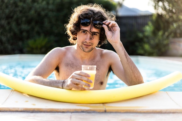 Cara usando óculos de sol se divertindo na beira da piscina bebendo suco de laranja