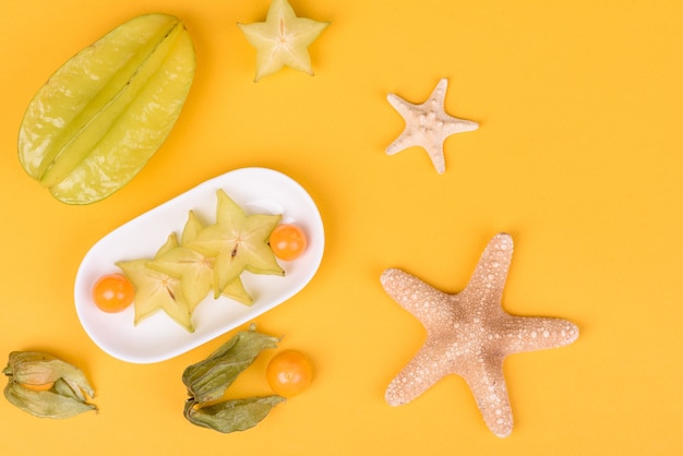 Carambol de frutas, accesorios de playa y follaje de una planta tropical sobre papel de colores. Fondo de verano con estrellas de mar y frutas.