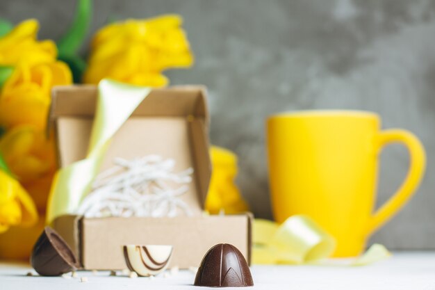 Caramelos de chocolate en caja artesanal, taza y ramo de tulipanes amarillos sobre superficie de madera blanca en pared gris. Copia espacio