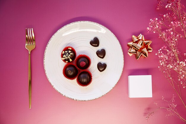 Caramelos de chocolate hechos a mano en forma de corazón en el plato con tenedor Caja blanca maqueta concepto de regalo creativo Vista superior Concepto de cena romántica Fondo degradado rosa