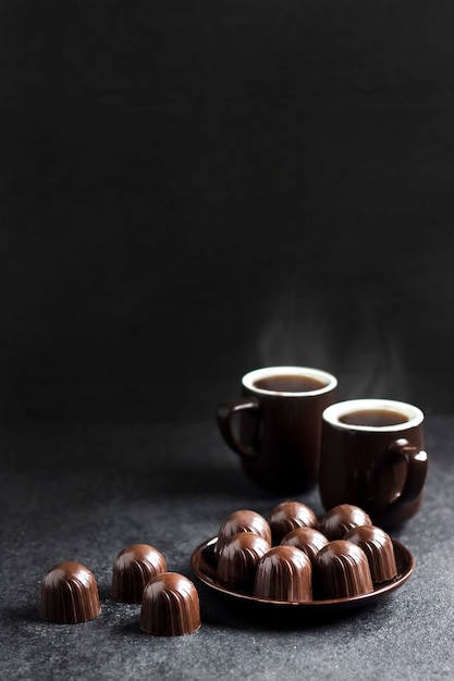 Caramelos de chocolate en un plato y dos tazas de café caliente sobre fondo negro con espacio de copia