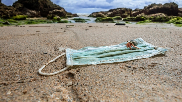 Caranguejo eremita caminhando sobre um lixo de máscaras médicas usadas na água do mar. Consequências ruins como poluição ou contaminação da natureza dos oceanos. Contaminação ambiental e costeira Covid 19