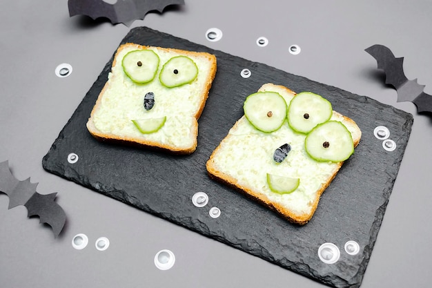 Caras de monstros engraçados no pão de torrada de sanduíche de halloween com pepinos de ricota de queijo de abacate verde macio no corte servindo boardbats em fundo cinzaCrianças criança café da manhã almoço comida