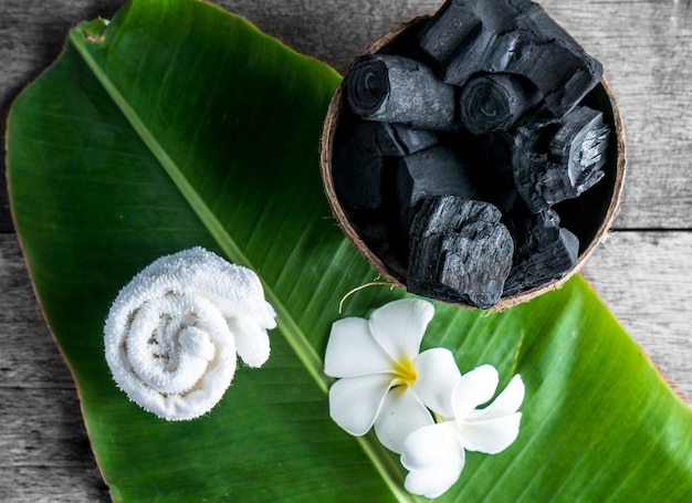 Carbón vegetal en cáscara de coco para spa en el fondo de madera con toalla blanca y flor en hoja de plátano