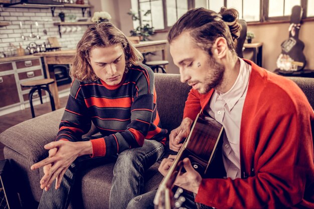 Cárdigan rojo. Músico barbudo con camisa blanca y chaqueta roja tocando la guitarra sentado cerca de su amigo