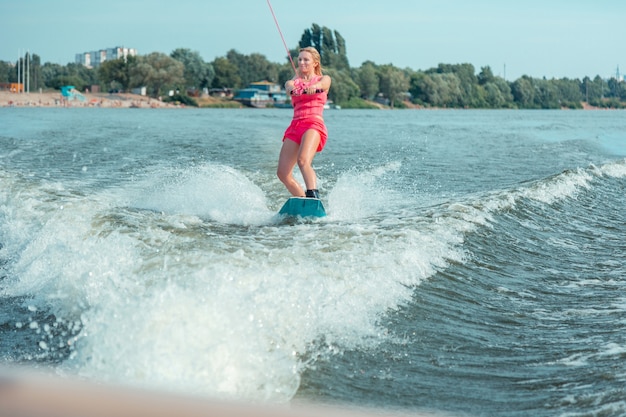 Cardio-Aktivität. Zufriedene schlanke blonde Frau in einer Schwimmweste steht selbstbewusst auf ihrem Wakeboard und genießt ihre Fahrt auf dem Fluss