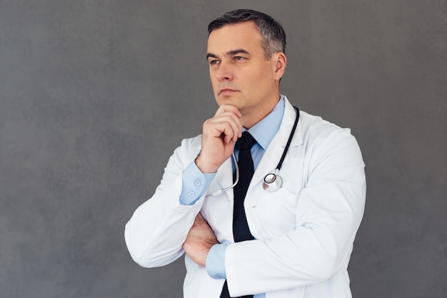 Cardiologista. Médico homem maduro mantendo a mão no queixo e olhando para longe enquanto fica de pé contra um fundo cinza
