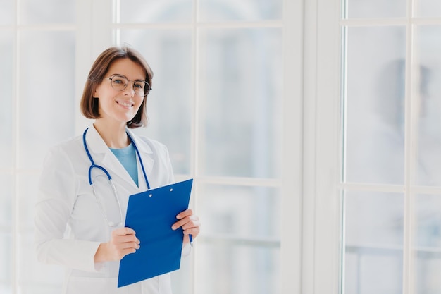 Foto la cardioterapeuta femenina usa anteojos redondos, abrigo médico blanco y fonendoscopio escribe con bolígrafo en soportes de portapapeles cerca de una gran ventana en su propia clínica, piensa en el caso y el tratamiento del paciente