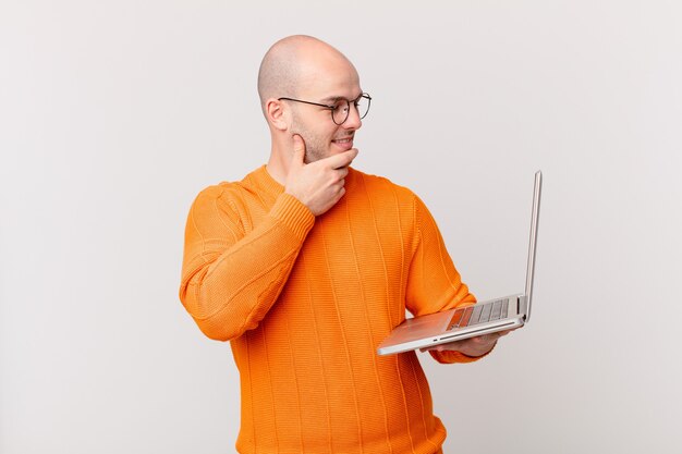 Careca com computador sorrindo com uma expressão feliz e confiante com a mão no queixo, pensando e olhando para o lado