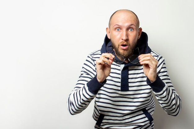 Careca Jovem com barba, um suéter com capuz tirou os óculos com um rosto surpreso em um branco isolado. Um gesto de surpresa, choque. Copie o espaço