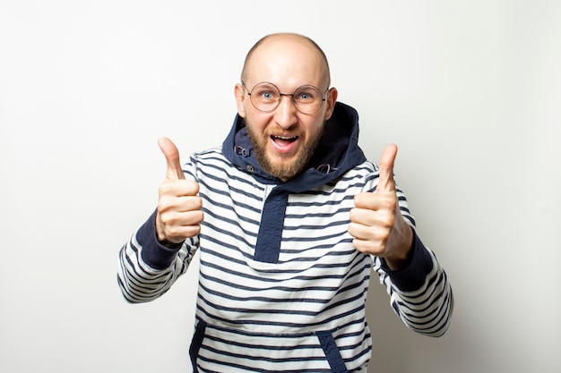 Careca jovem com uma barba de óculos, um suéter com capuz, mostra ao espectador um gesto com o polegar sobre um branco isolado. gesto está tudo bem, ok