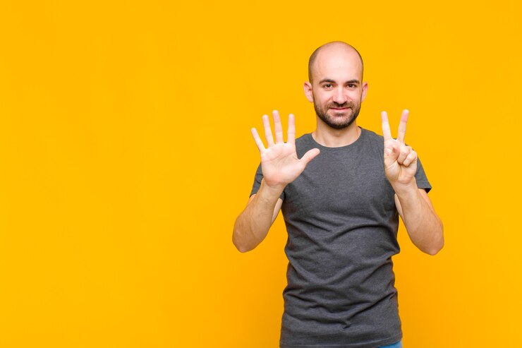 Foto careca sorrindo e parecendo amigável, mostrando o número sete ou sétimo com a mão para a frente, em contagem regressiva