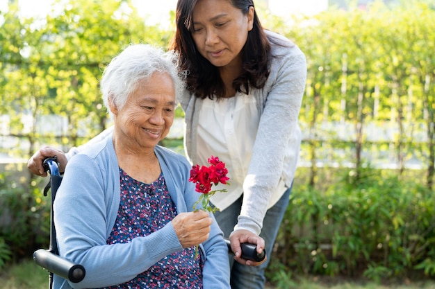 Caregver ayuda a una anciana asiática con una sonrisa de flor de rosa roja y feliz en el jardín soleado