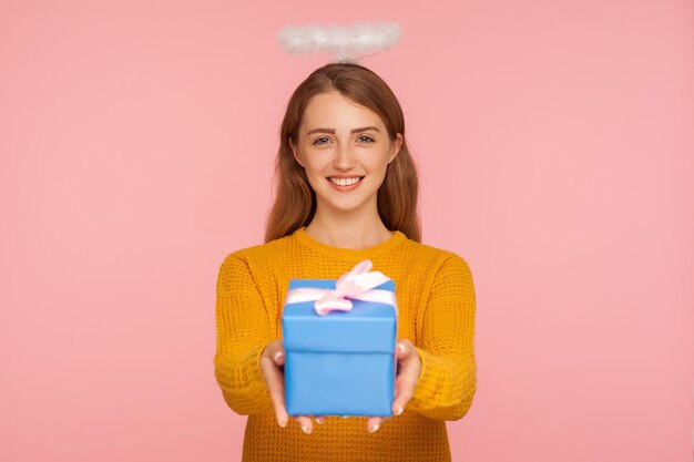 Caridade e doações. Retrato de menina ruiva generosa angelical com halo sobre a cabeça dando caixa de presente e sorrindo para a câmera, oferecendo presente nas férias. tiro de estúdio interior isolado no fundo rosa