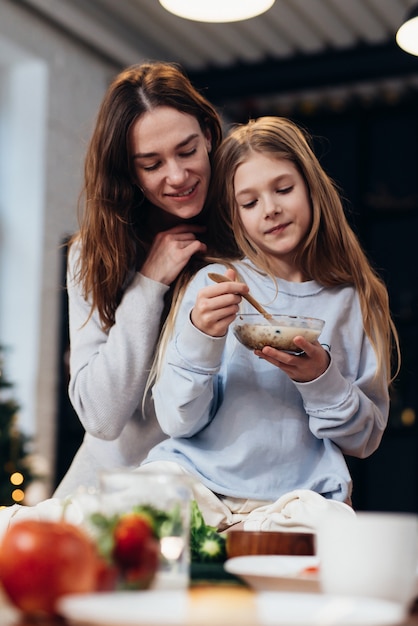 Carinhosa, abraçando a mãe ao lado de sua filha comendo granola.