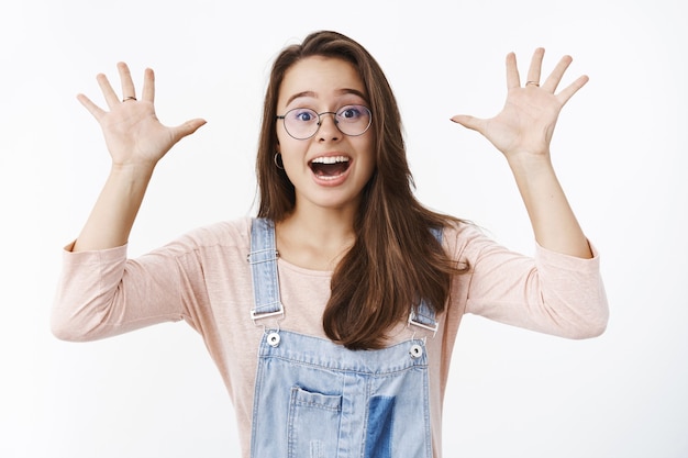 Carismática joven sorprendida y emocionada feliz linda mujer con gafas y overoles de mezclilla agitando las manos levantadas con asombro gritando sí de emoción y felicidad, animando, celebrando la victoria.