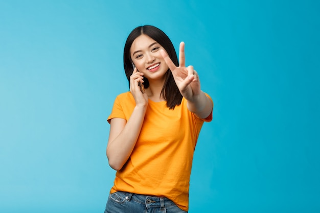 Carismática linda chica comparte noticias positivas hablando teléfono inteligente muestra paz, signo de victoria sonriendo satisfecho, fondo azul de pie recibe una agradable llamada telefónica, logra un trato a través de una llamada telefónica.