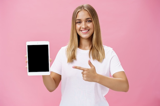 Carismática mujer joven con cabello rubio, piel bronceada y sin maquillaje en camiseta blanca apuntando a la pantalla del dispositivo y sonriendo amigablemente a la cámara sobre una pared rosa