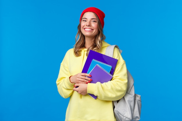 Carismática mujer sonriente con gorro rojo y sudadera amarilla, caminando a la universidad, llevar mochila y cuadernos, documentos para las clases, mirar con alegría, estudiar prepararse para los exámenes para ingresar a la universidad