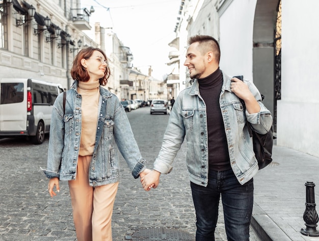 Carismática pareja moderna de turistas amorosos en chaquetas de mezclilla pasean tomados de la mano por la calle urbana