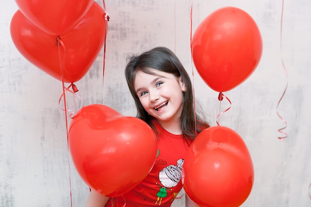 Carita de niña sonriente divertida de cerca entre globos rojos en forma de corazón