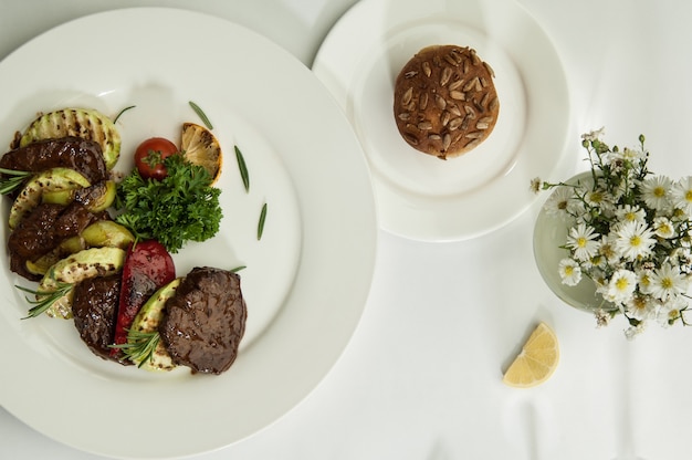 Carne asada con rodajas de limón sobre una superficie blanca con la sombra de las copas de vino