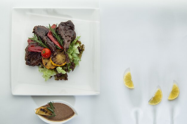 Carne assada com rodelas de limão em uma superfície branca com a sombra das taças de vinho