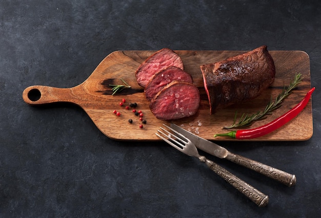 Carne assada em uma tábua de madeira com alecrim e pimenta em uma vista superior de fundo preto concreto