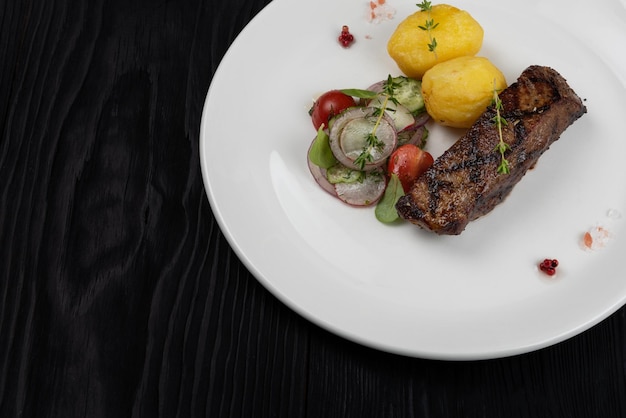 Carne de bistec de falda de ternera a la parrilla con patatas y verduras en un plato blanco sobre fondo negro de madera